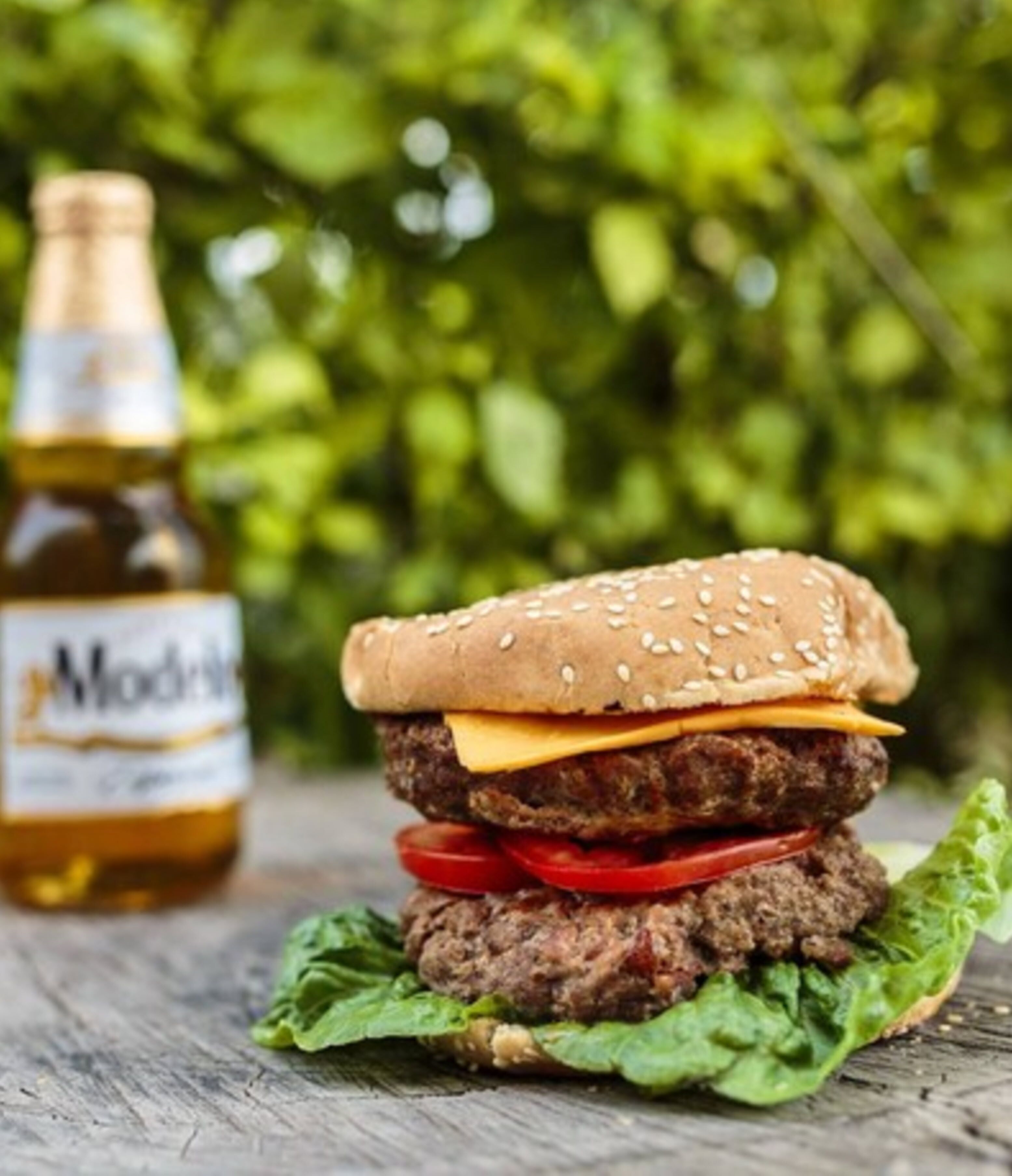 Una hamburguesa con lechuga, tomate y queso, y una botella de cerveza ModClarke al fondo.