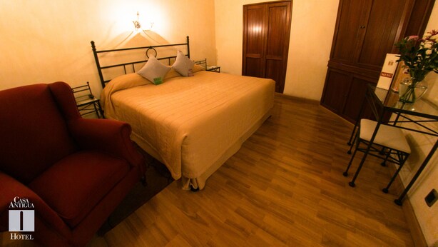 A bedroom with a bed, a red armchair, a wooden wardrobe, and a small table with a vase.