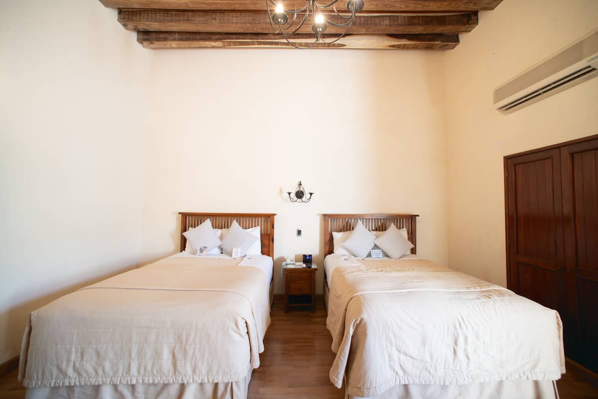 Quarto com duas camas, cabeceiras de madeira, uma mesinha de cabeceira pequena, luminária fixada na parede e guarda-roupa de madeira.