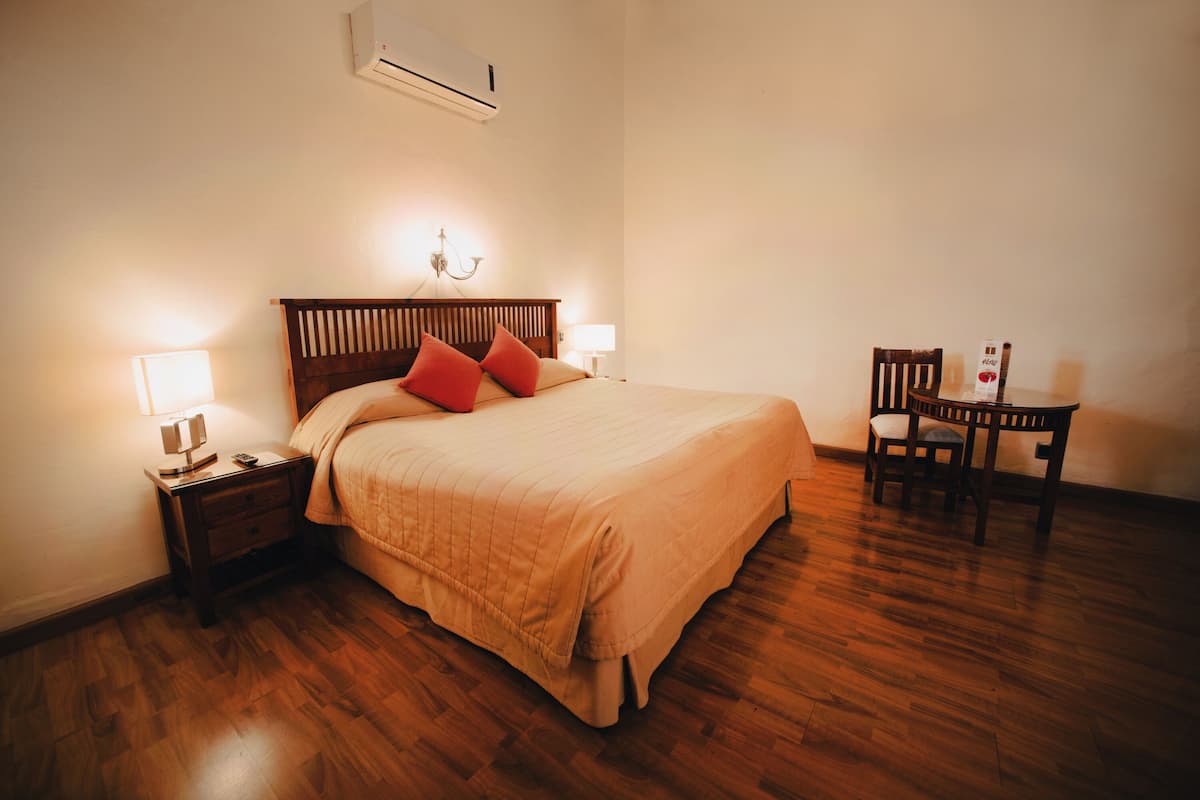 Cama bem arrumada com cabeceira de madeira, mesa de cabeceira com abajur e uma cadeira.