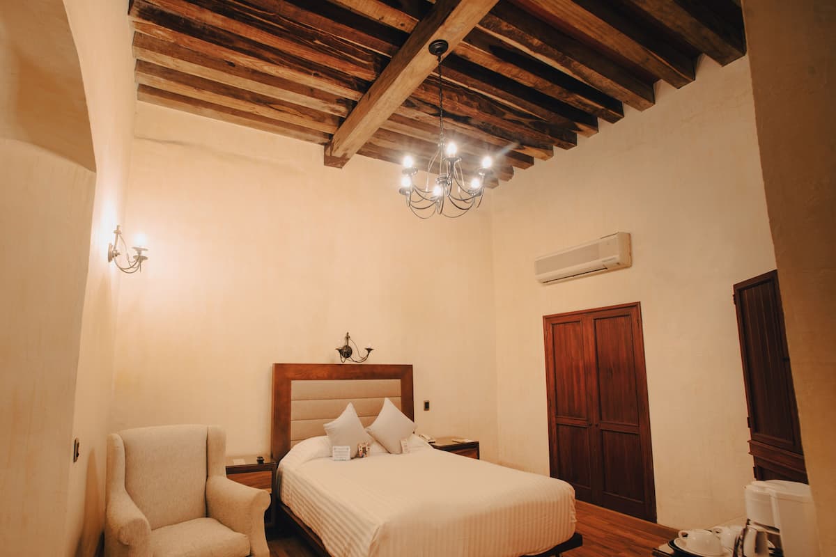 Quarto com teto de madeira, cama com cabeceira de madeira, uma cadeira, mesa de cabeceira e cafeteira.