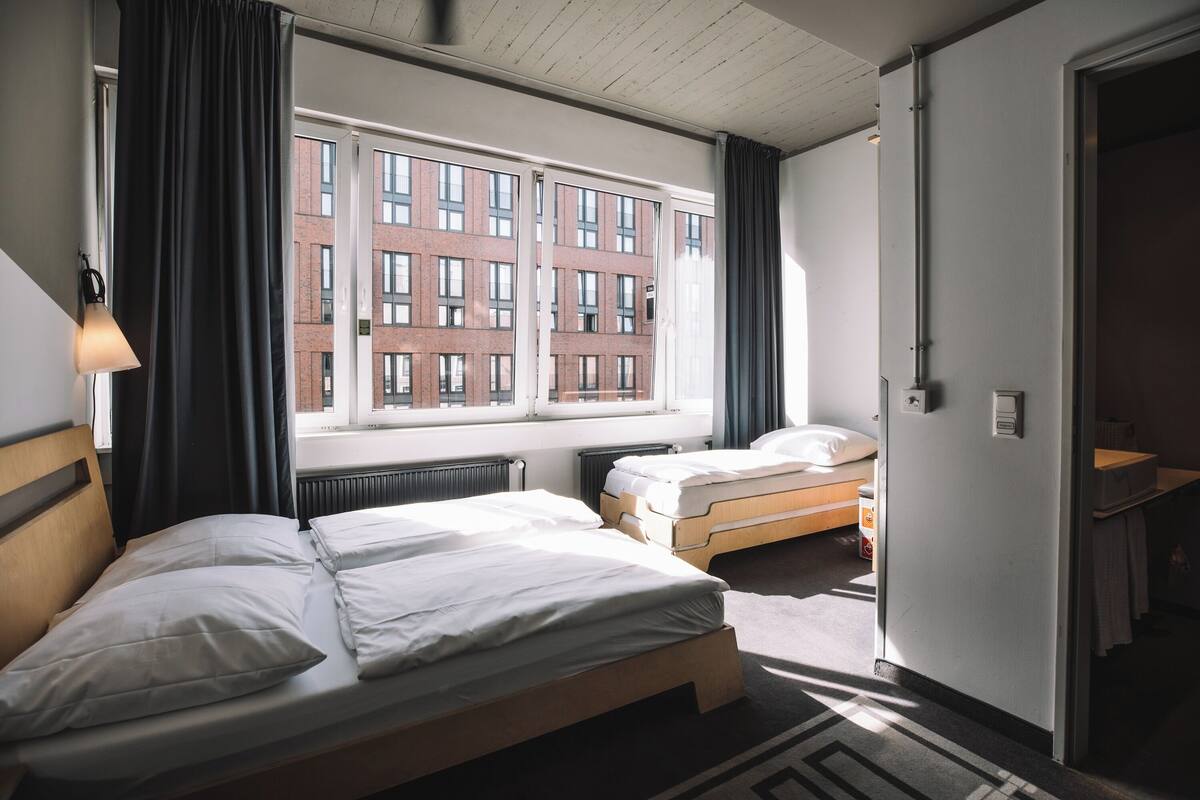 A hotel room with two beds, a window with a view of a brick building, a radiator, and a door leading to a bathroom.