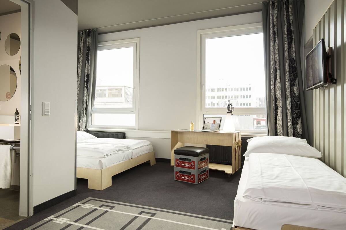 A hotel room with two beds, a desk with a laptop, a radiator, and a window with curtains.
