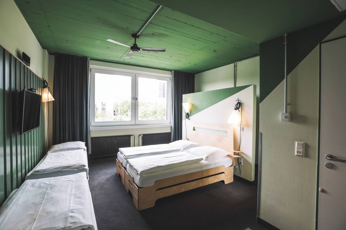 A room with two single beds, a ceiling fan, a window with curtains, a bedside lamp, and a door with a handle.