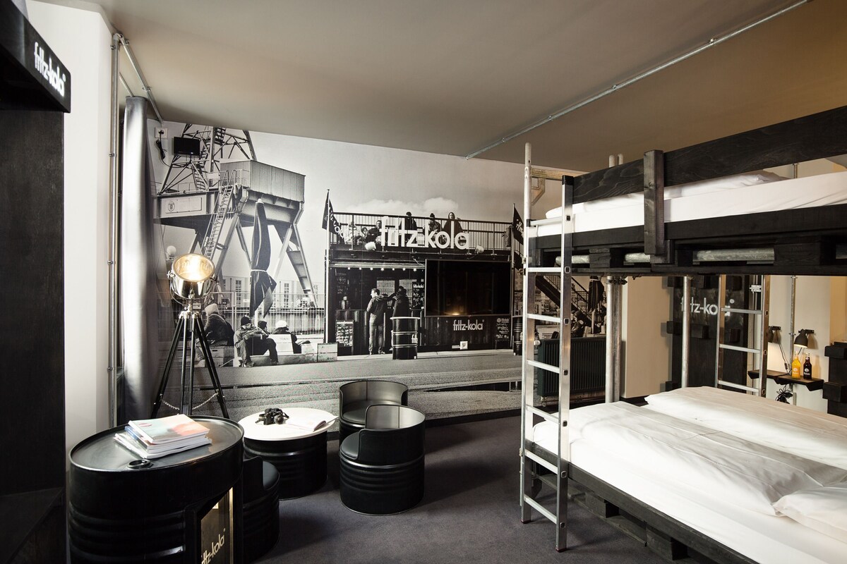 A modern hotel room with a bunk bed, a desk with a lamp, a seating area with stools, and a mural of a cityscape.