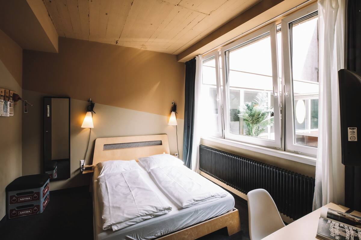 A hotel room with a bed, bedside lamps, a radiator, a window with curtains, a desk, and a chair.