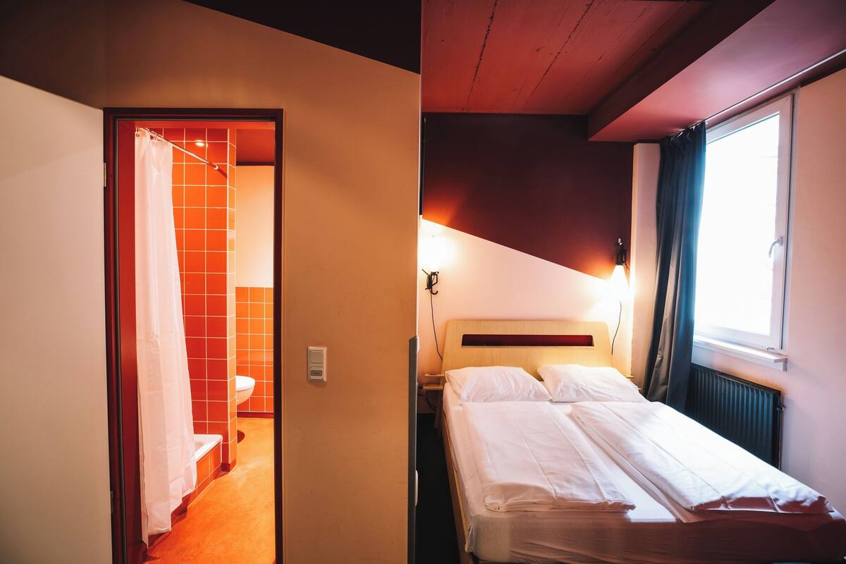 A hotel room with a bed, a window with curtains, a shower area, and a wall-mounted light.