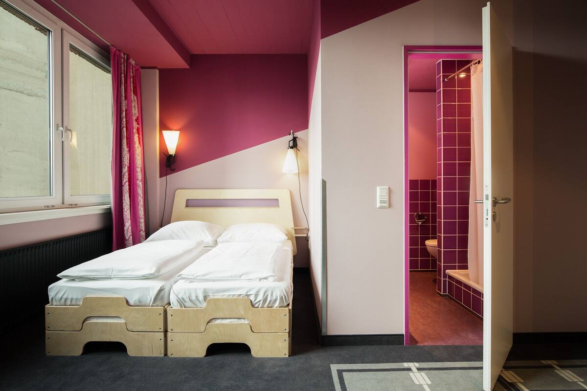 A room with a bed, a window with curtains, a wall-mounted light, and a door leading to a bathroom.