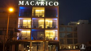 Un hotel de varios pisos llamado MACARICO con señalización iluminada, un cartel de playa visible y un diseño arquitectónico moderno.