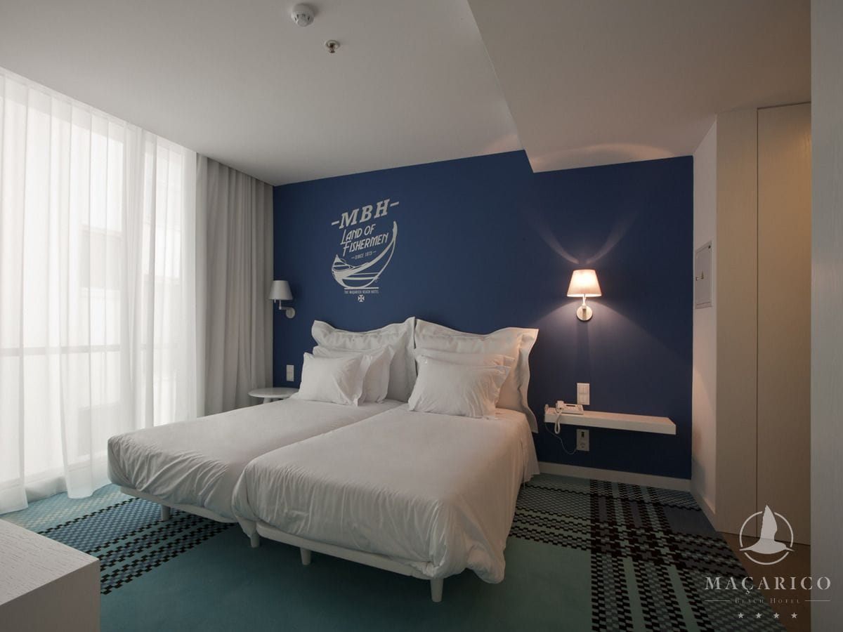 A hotel room with a bed, bedside tables, a lamp, and a wall with a logo.