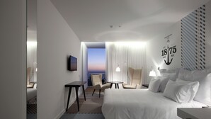 Habitación de hotel con una cama grande, dos sillones, una mesita y un televisor. Se ve un atardecer a través de la ventana.