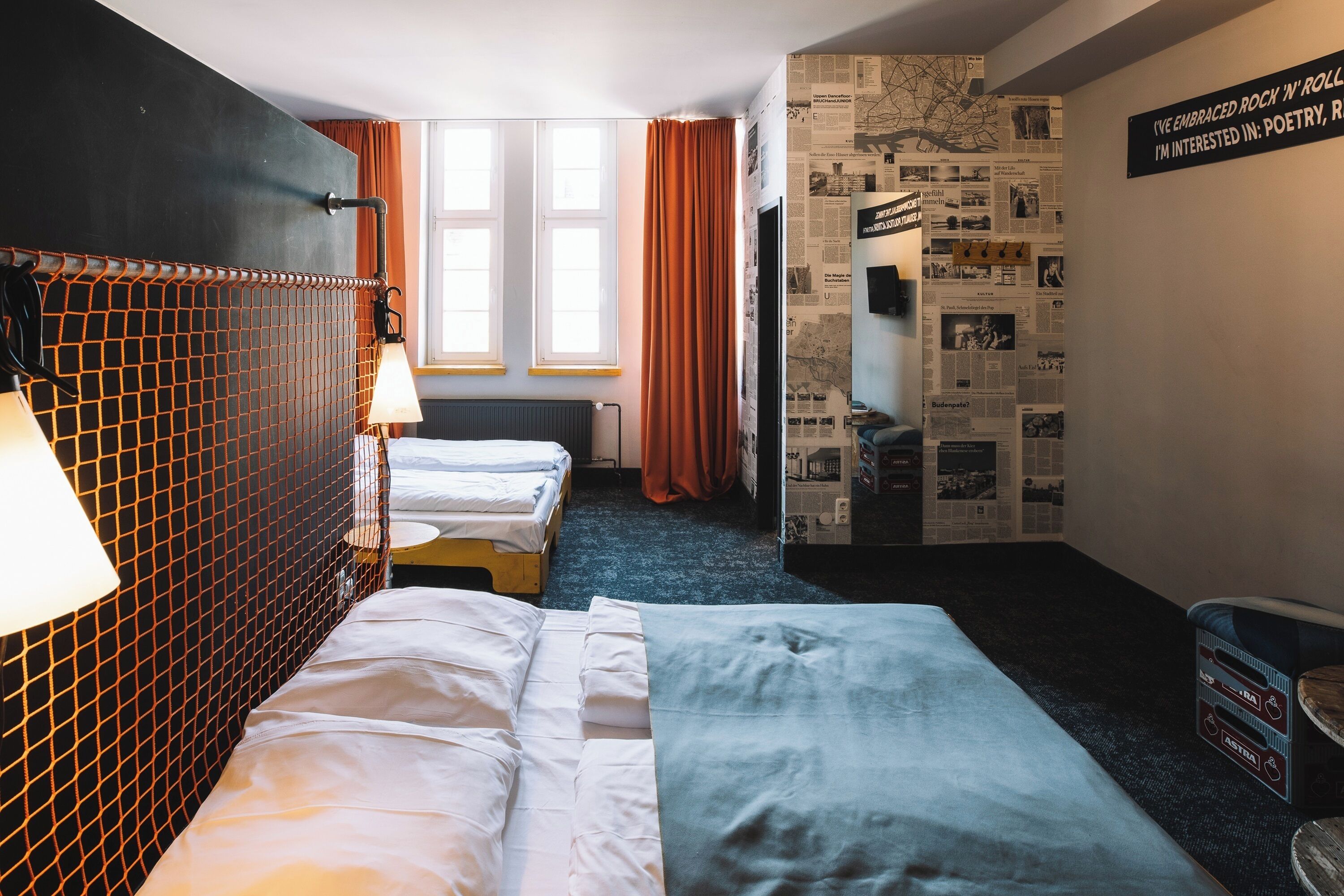 A hotel room with two beds, a large window with curtains, a wall with newspaper clippings, and a sign about the guest's interests.