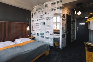Une chambre d’hôtel avec un lit, une tête de lit en bois, un mur recouvert de coupures de journaux et une porte donnant sur une autre pièce.