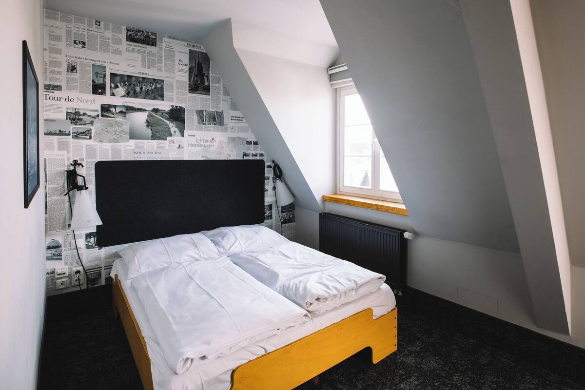 A bedroom with a bed, a window, and a wall covered with newspaper clippings.
