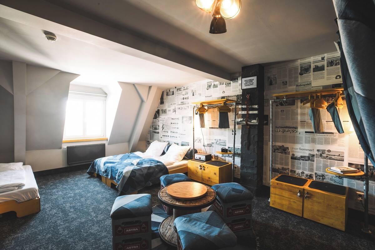 A compact hotel room with a sloped ceiling, a bed with blue bedding, a small wooden table, and a wall adorned with newspaper clippings.