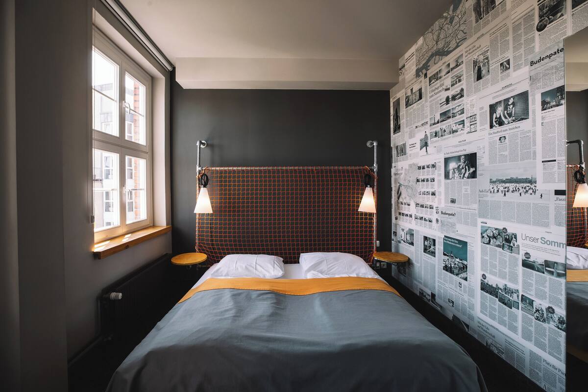 A hotel room with a large bed, two bedside tables, a window, and a wall covered with newspaper clippings.