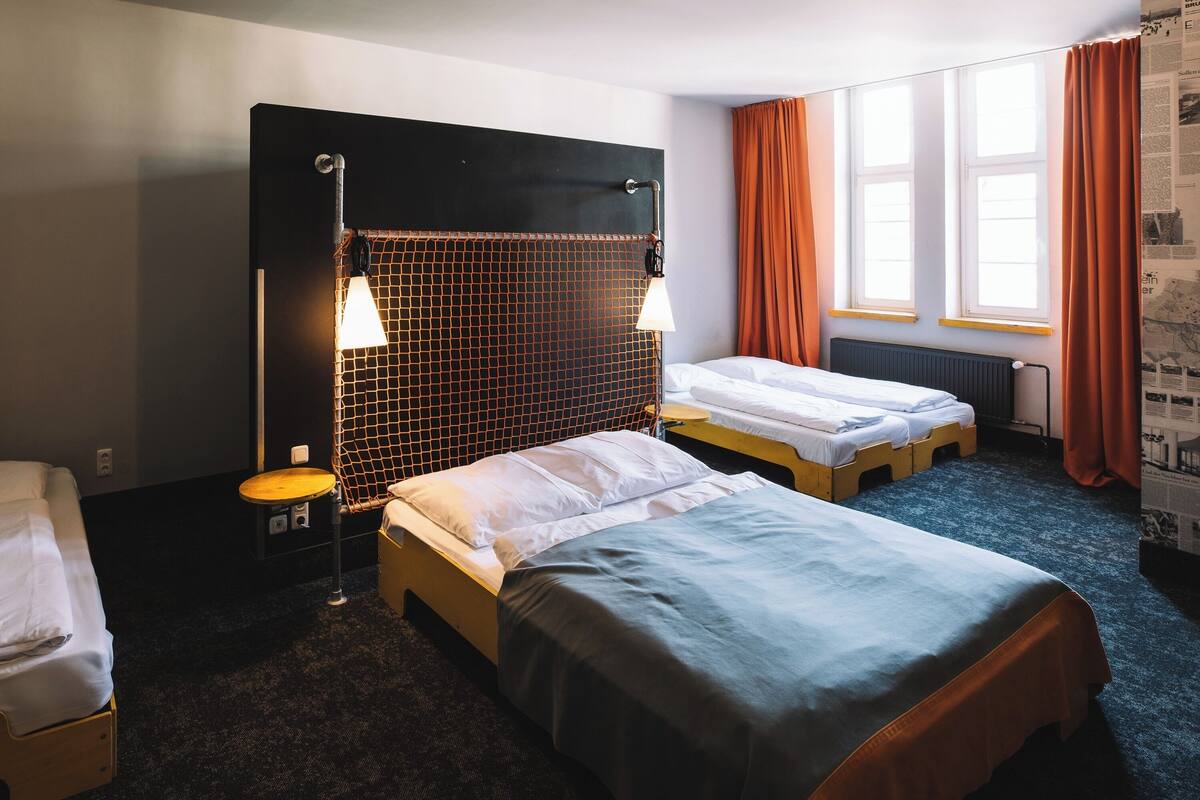 A hotel room with two single beds, a small table, a chair, a desk, and a window with curtains.