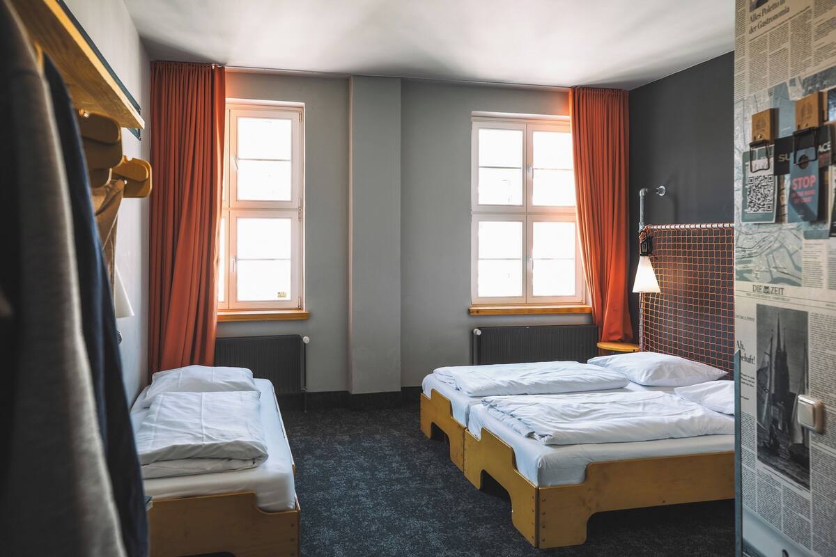 A room with two single beds, a radiator, a window with curtains, and a wall with a poster.