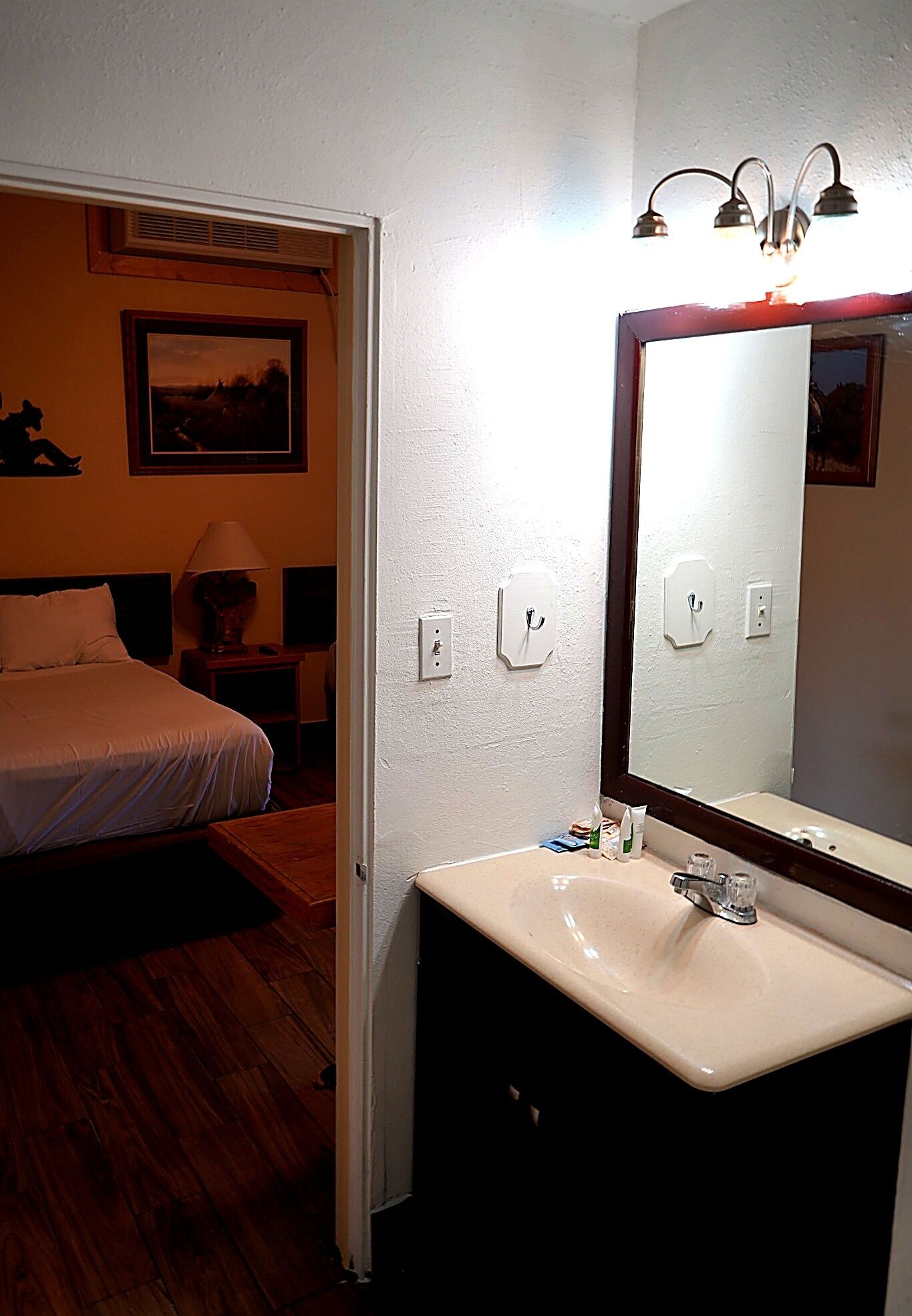 A hotel room with a bed, a bedside table with a lamp, a bathroom with a sink and mirror, and a painting on the wall.