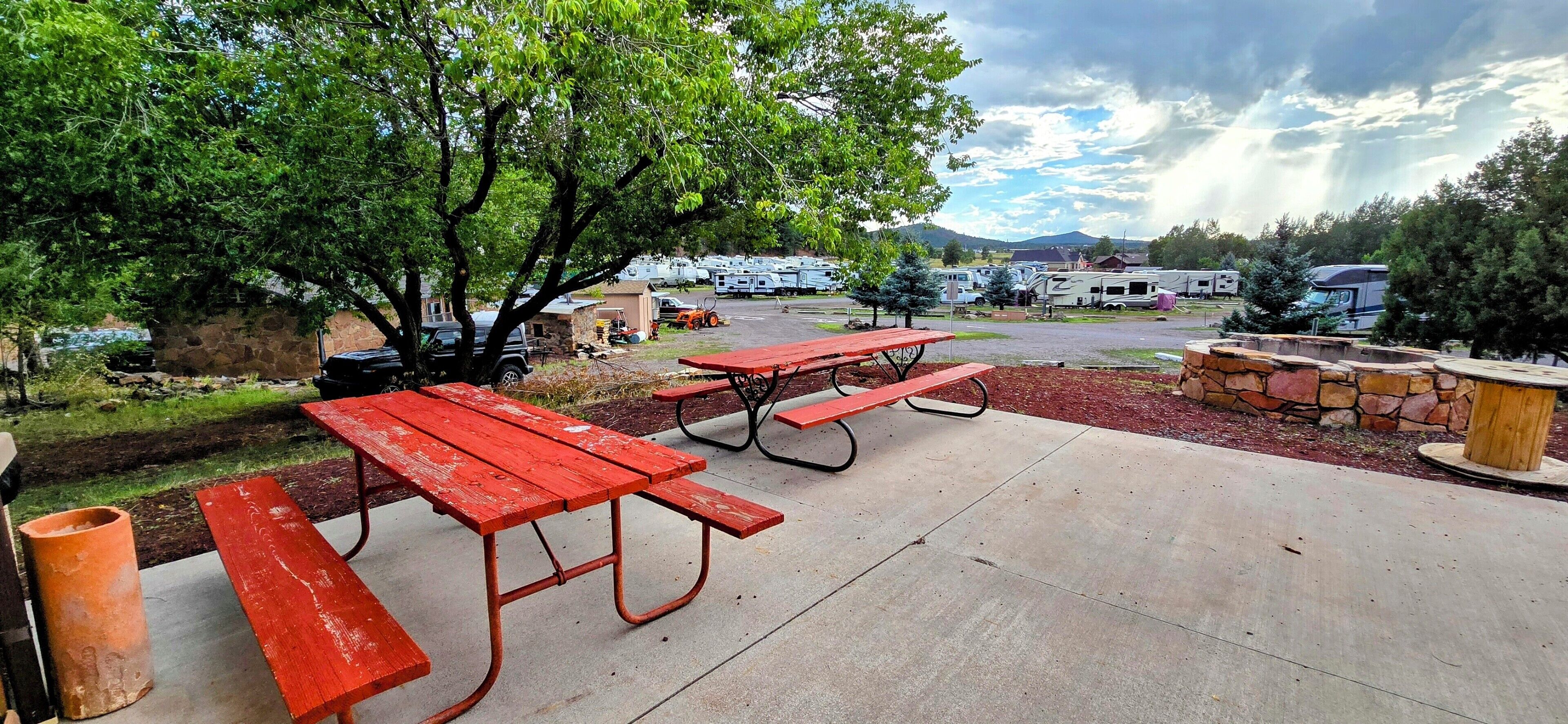 Un área de picnic con bancos rojos y un hogar de piedra, rodeado de árboles y vehículos recreativos al fondo.