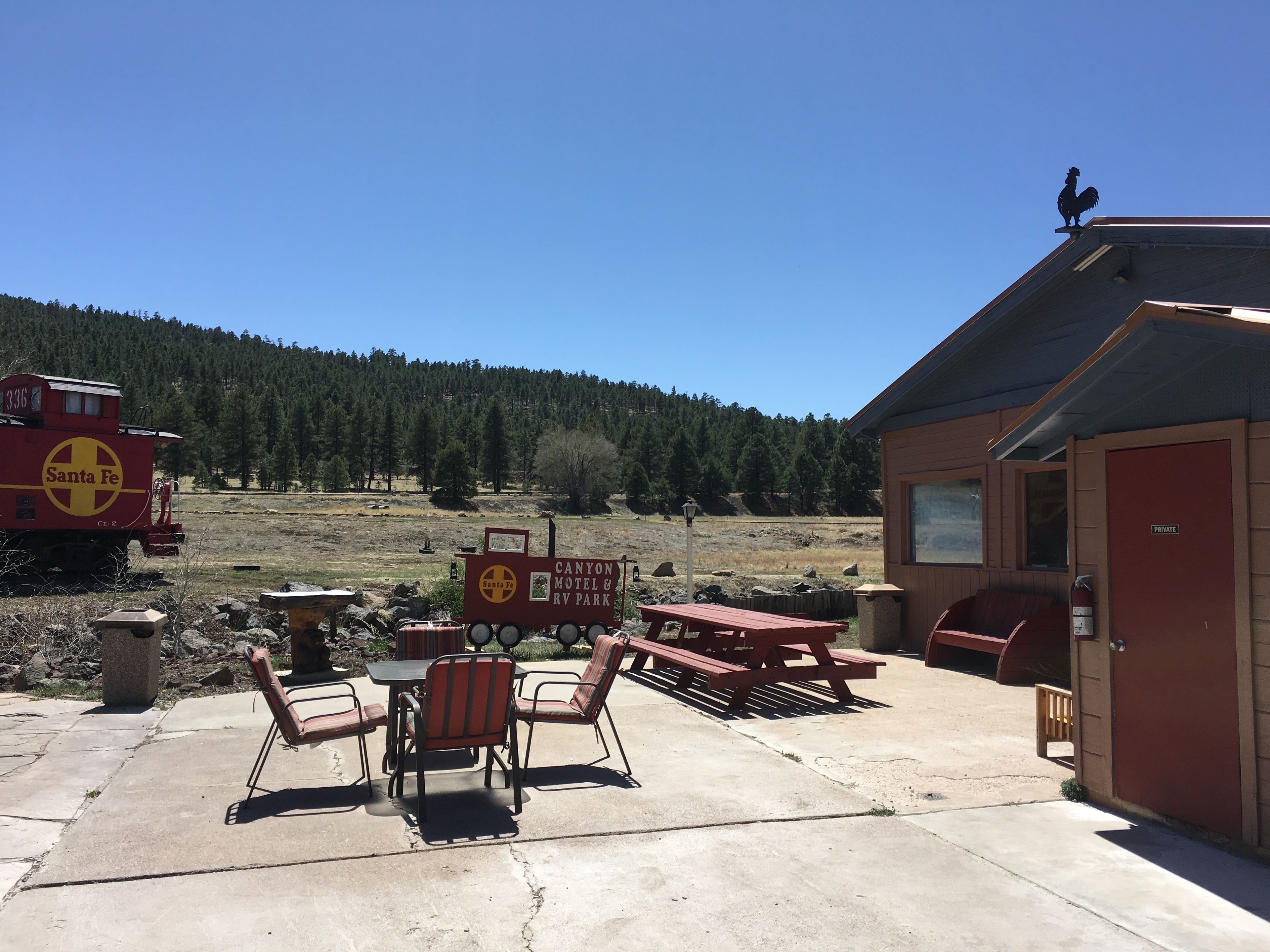Un motel rústico con una mesa y sillas de picnic, un edificio rojo, y un tren al fondo.