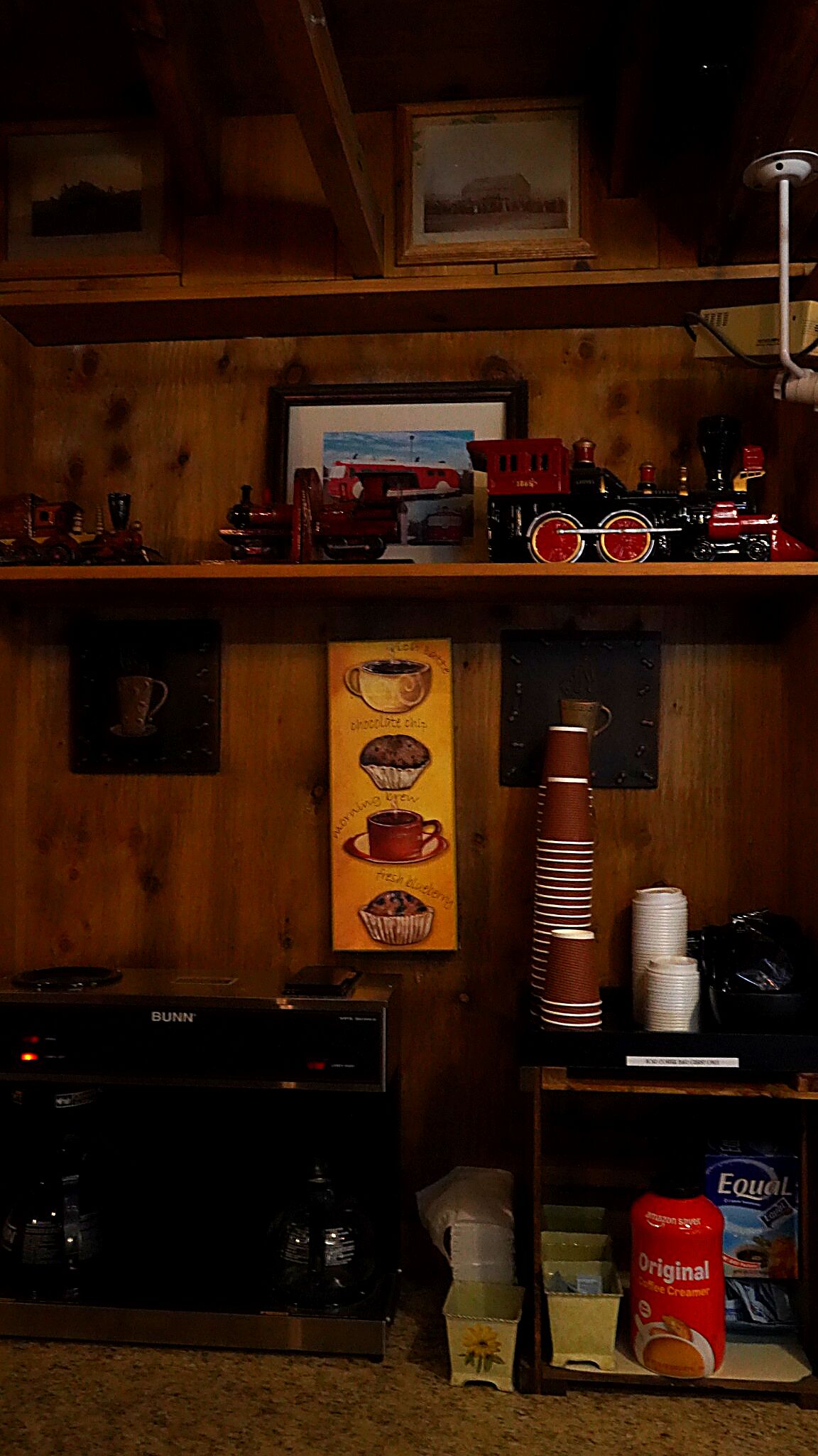 Une pièce chaleureuse aux murs en bois, avec une machine à expresso de la marque Bunn, une table basse et une étagère où sont disposés divers objets, notamment un tableau encadré, une illustration représentant une tasse de café et une pile d’assiettes.
