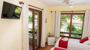 Habitación de hotel con televisión, dos camas con ropa de cama roja y blanca, una mesita con una botella y aperitivos, y vista a zonas verdes por las ventanas.
