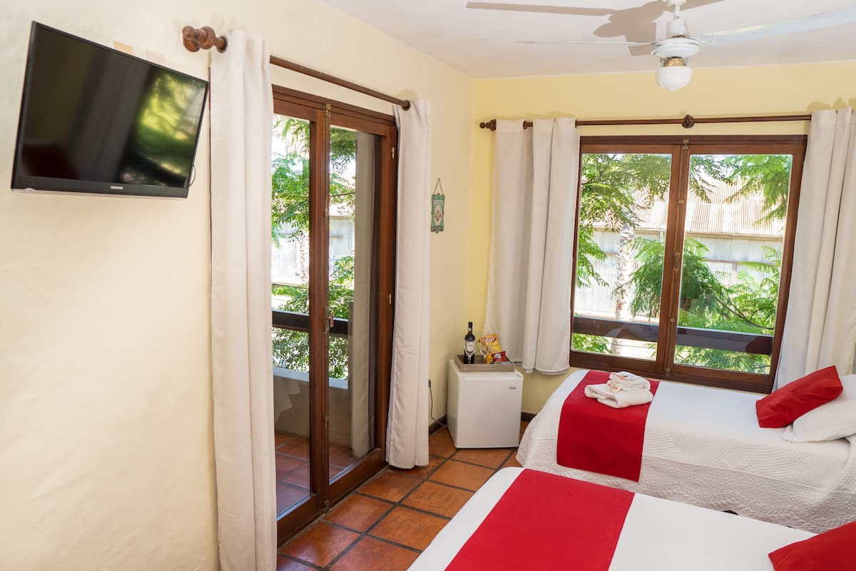 A hotel room with a TV, two beds with red and white bedding, a small table with a bottle and snacks, and a view of greenery through the windows.