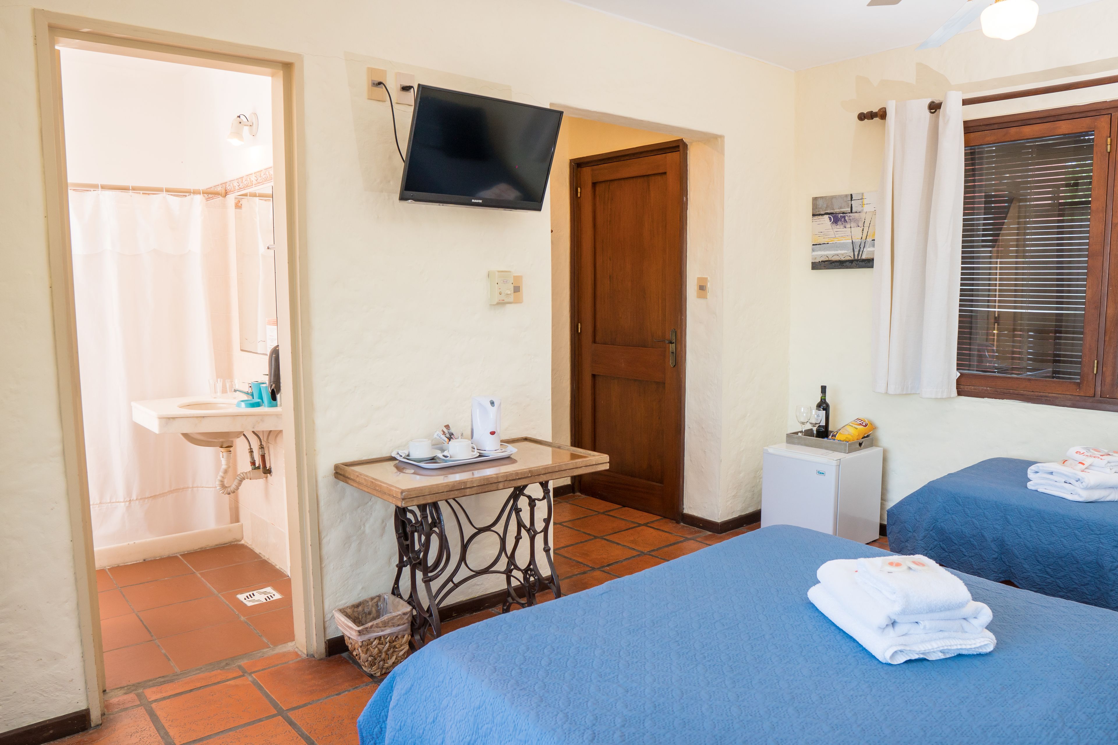 A hotel room with a bed, a small table with a kettle, a TV, a window with curtains, and a door.