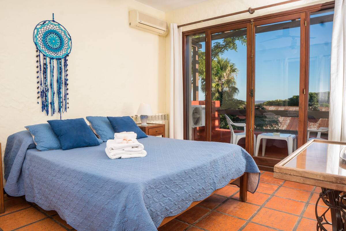 A bedroom with a bed, a bedside table, a dreamcatcher, and a view of the outdoors through a sliding glass door.