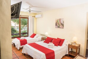 Une chambre d’hôtel avec deux lits, une literie rouge et blanche, un ventilateur de plafond, un appareil de climatisation, une table de chevet avec une lampe et une fenêtre donnant sur un paysage verdoyant.