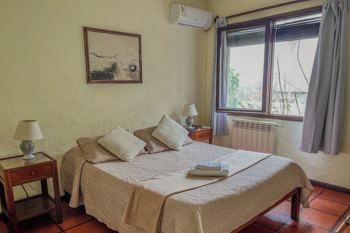 A neatly arranged bedroom with a bed, bedside lamps, a radiator, and a window with curtains.