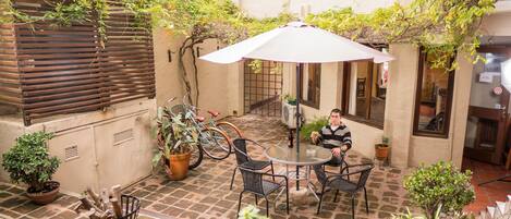 A courtyard with a round table, chairs, and a large umbrella. There are potted plants and a bicycle in the background.