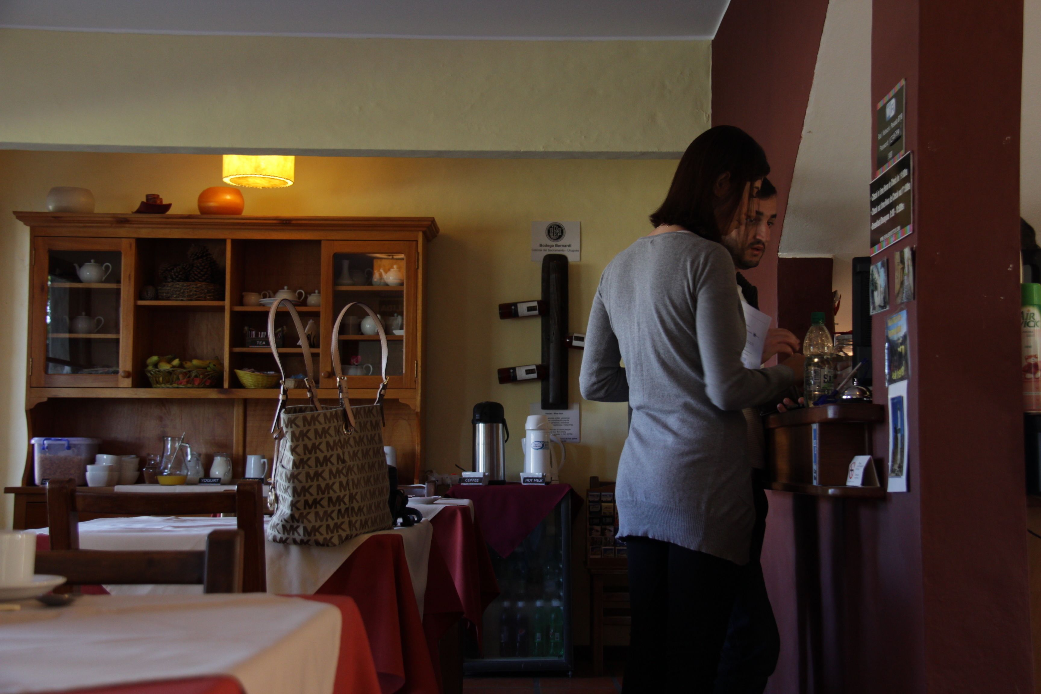 A person standing by a counter in a room with a wooden cabinet, a table with a red tablecloth, and various kitchen items.
