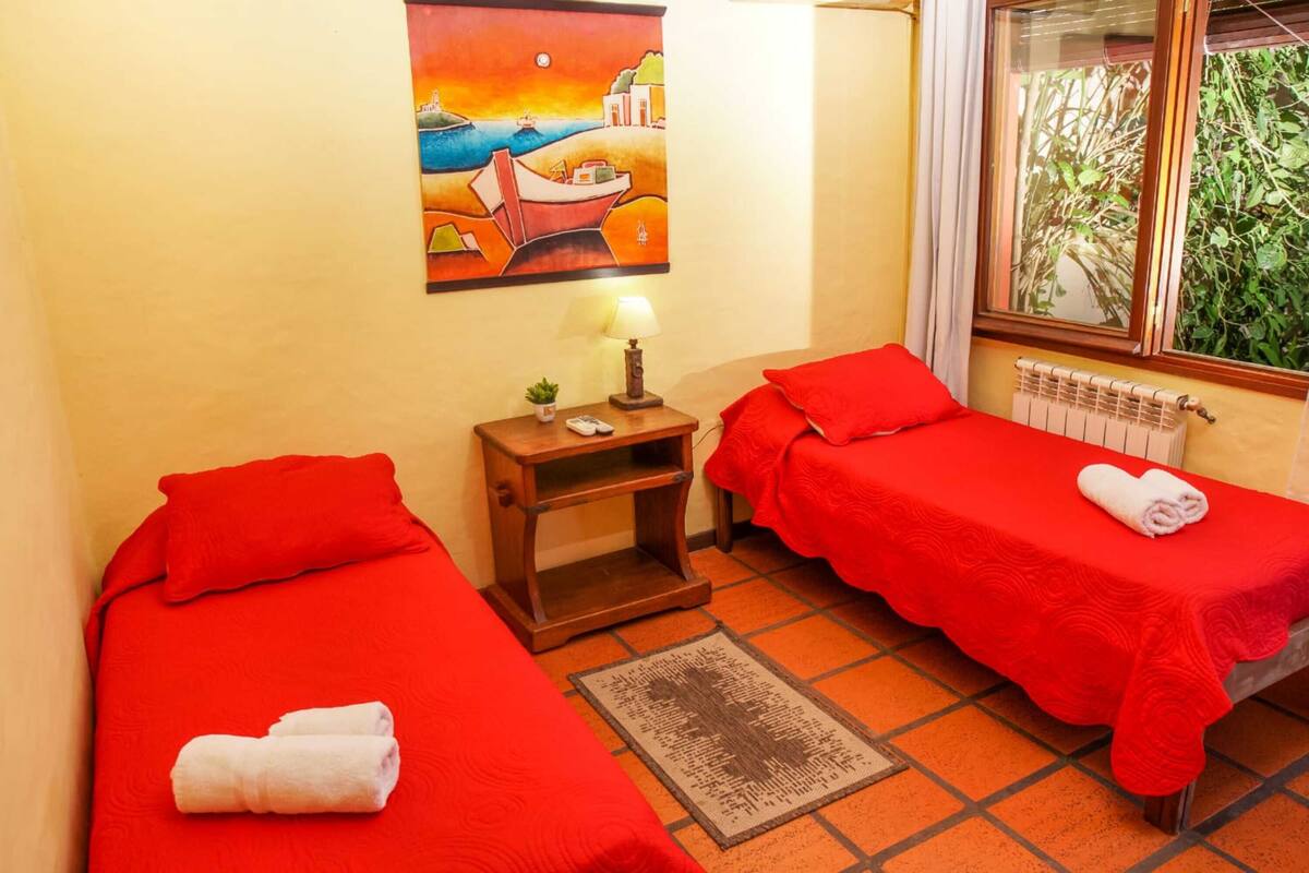 A room with two single beds, each with a red bedspread and a rolled white towel. There is a painting of a coastal scene on the wall, a small table with a lamp, and a window with a view of greenery.
