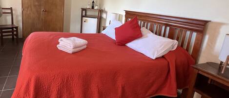 Una cama bien tendida con una colcha roja, almohadas blancas y una toalla doblada.