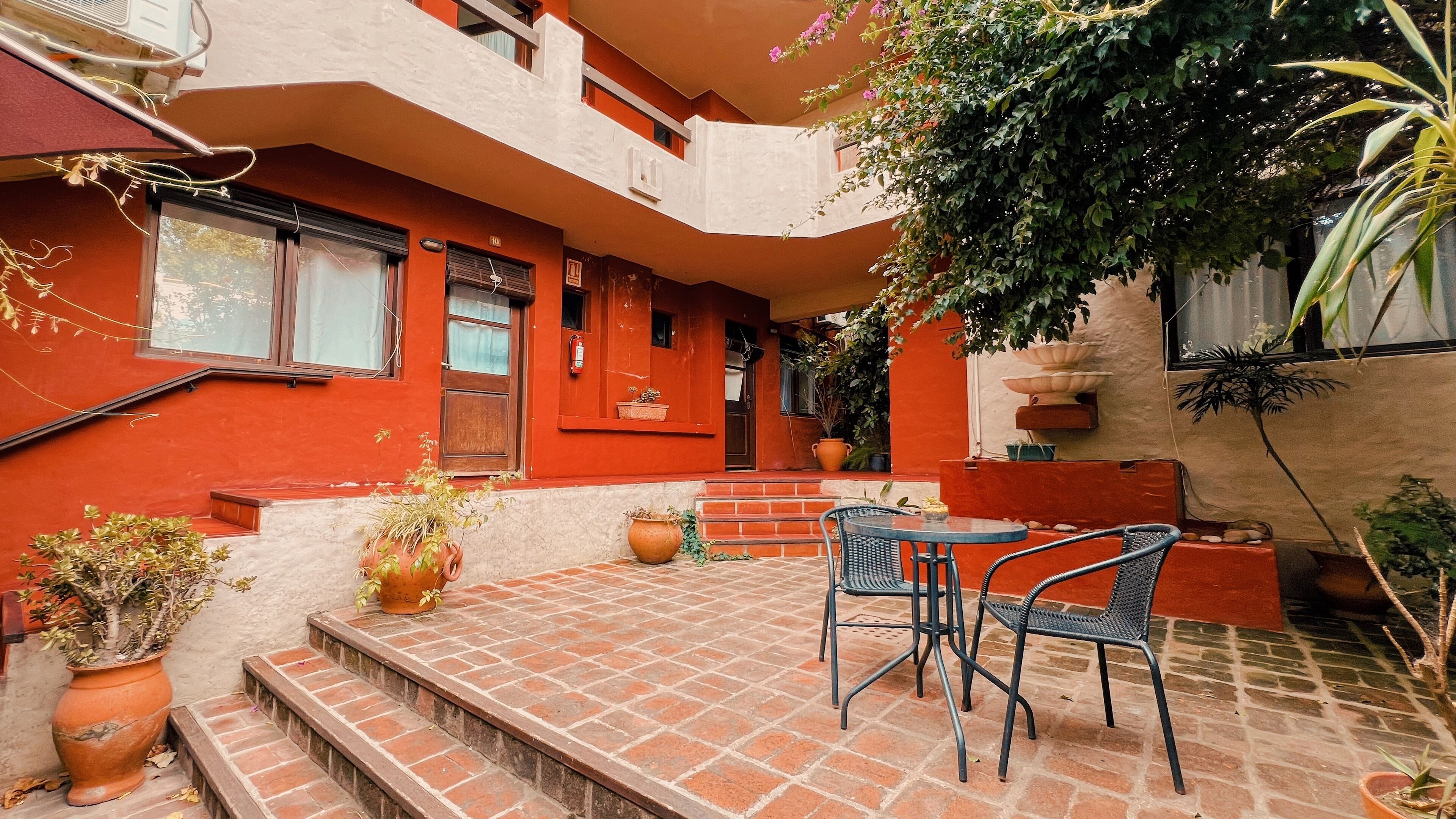 A red building with a patio area featuring a table and chairs, potted plants, and a staircase leading to the entrance.