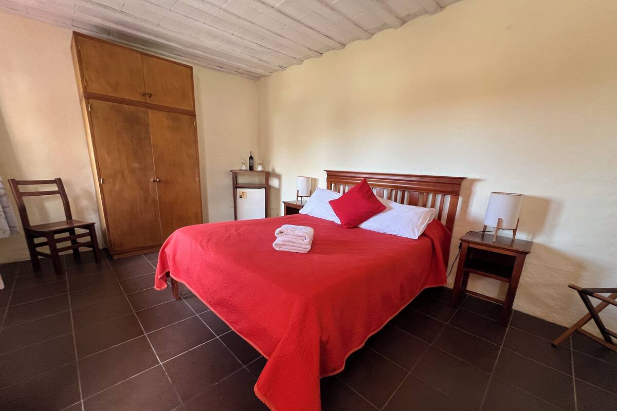 A neatly made bed with a red bedspread, a wooden headboard, and a white pillow. There is a wooden nightstand with a lamp beside the bed. A wooden chair is placed against the wall, and a wooden wardrobe is visible in the corner.