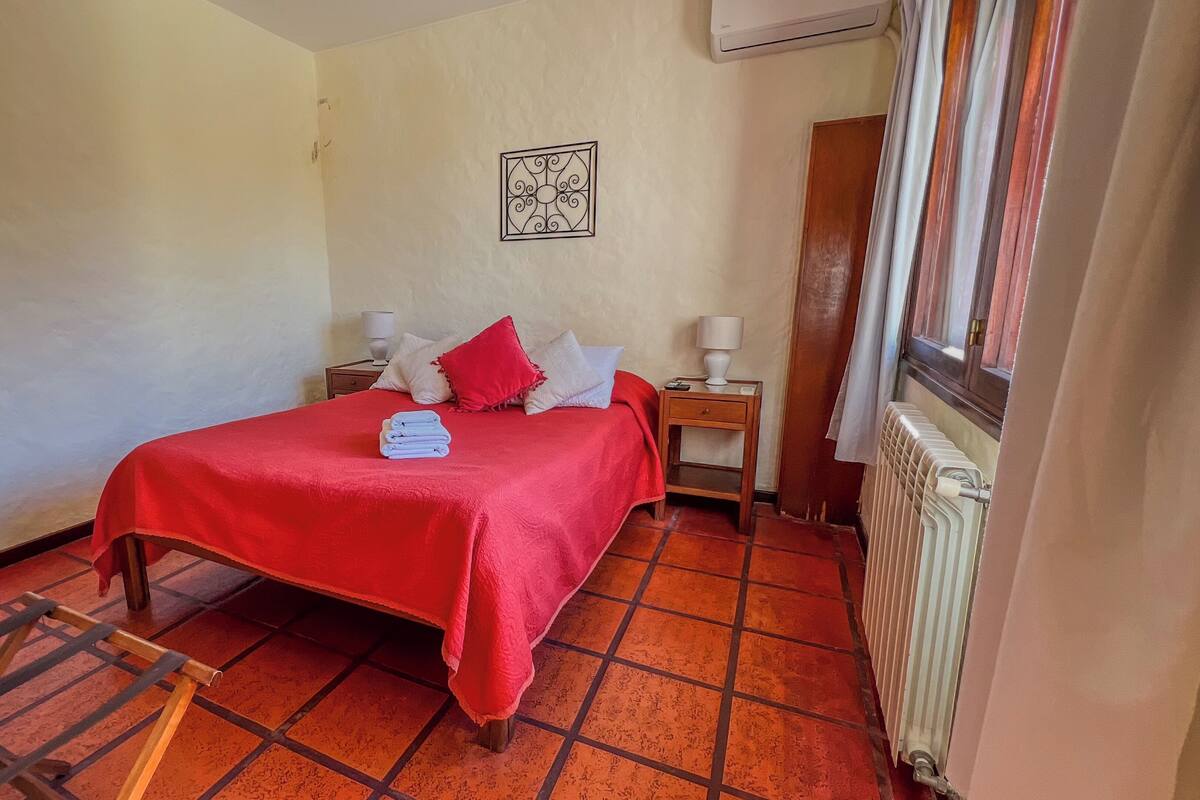 A small, neatly arranged bedroom with a red bedspread, a radiator, and a window with curtains.