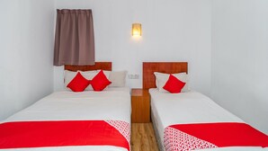 A hotel room with two beds, each with a wooden headboard, white bedding, and red accents. A wall-mounted light fixture is visible above the beds.