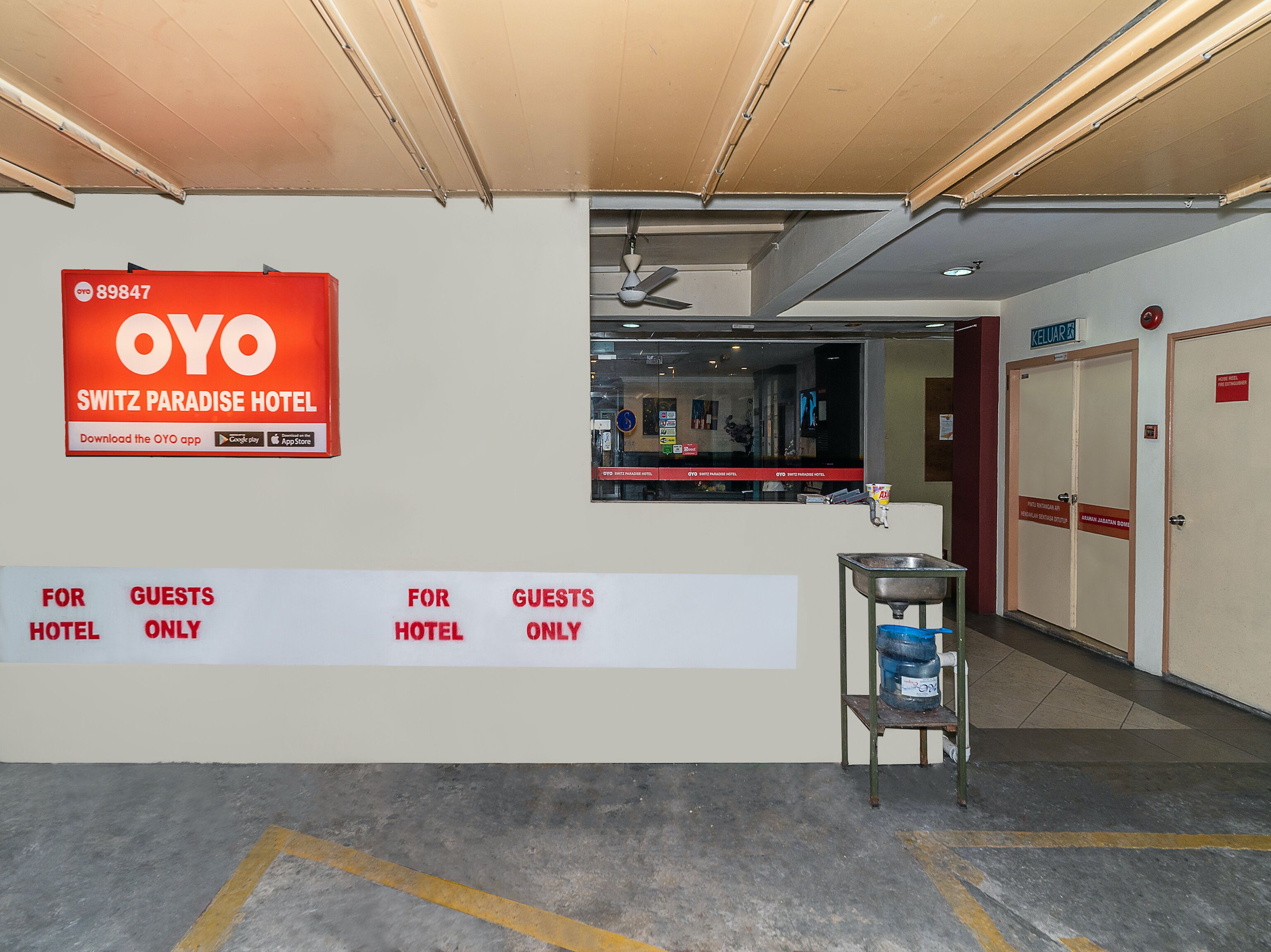 A hotel reception area with a sign for OYO SWITZ PARADISE HOTEL, a water dispenser, and a sign indicating guest-only areas.