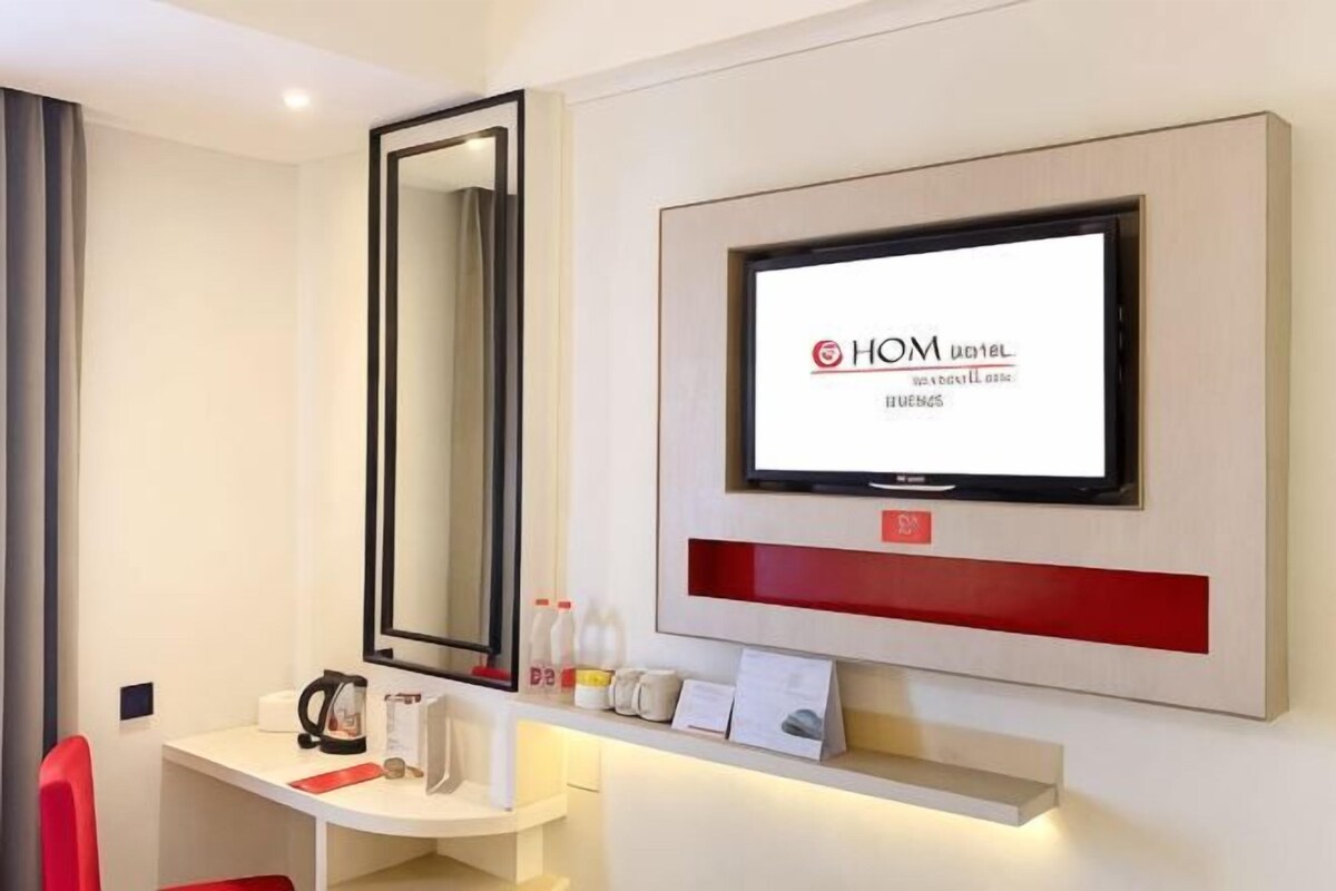 A hotel room with a TV mounted on the wall, a mirror, a desk with a red chair, and a shelf with toiletries.