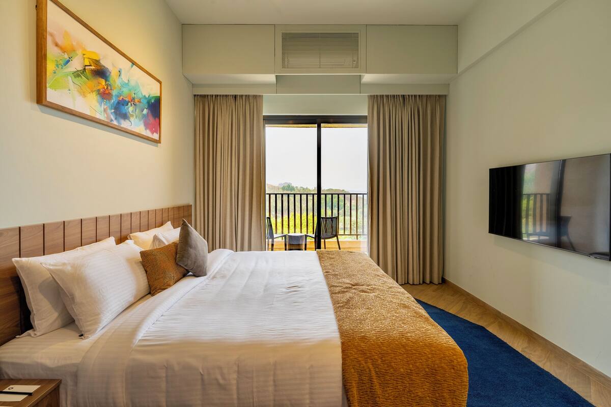 A hotel room with a large bed, a flat-screen TV, a painting on the wall, and a balcony with curtains.