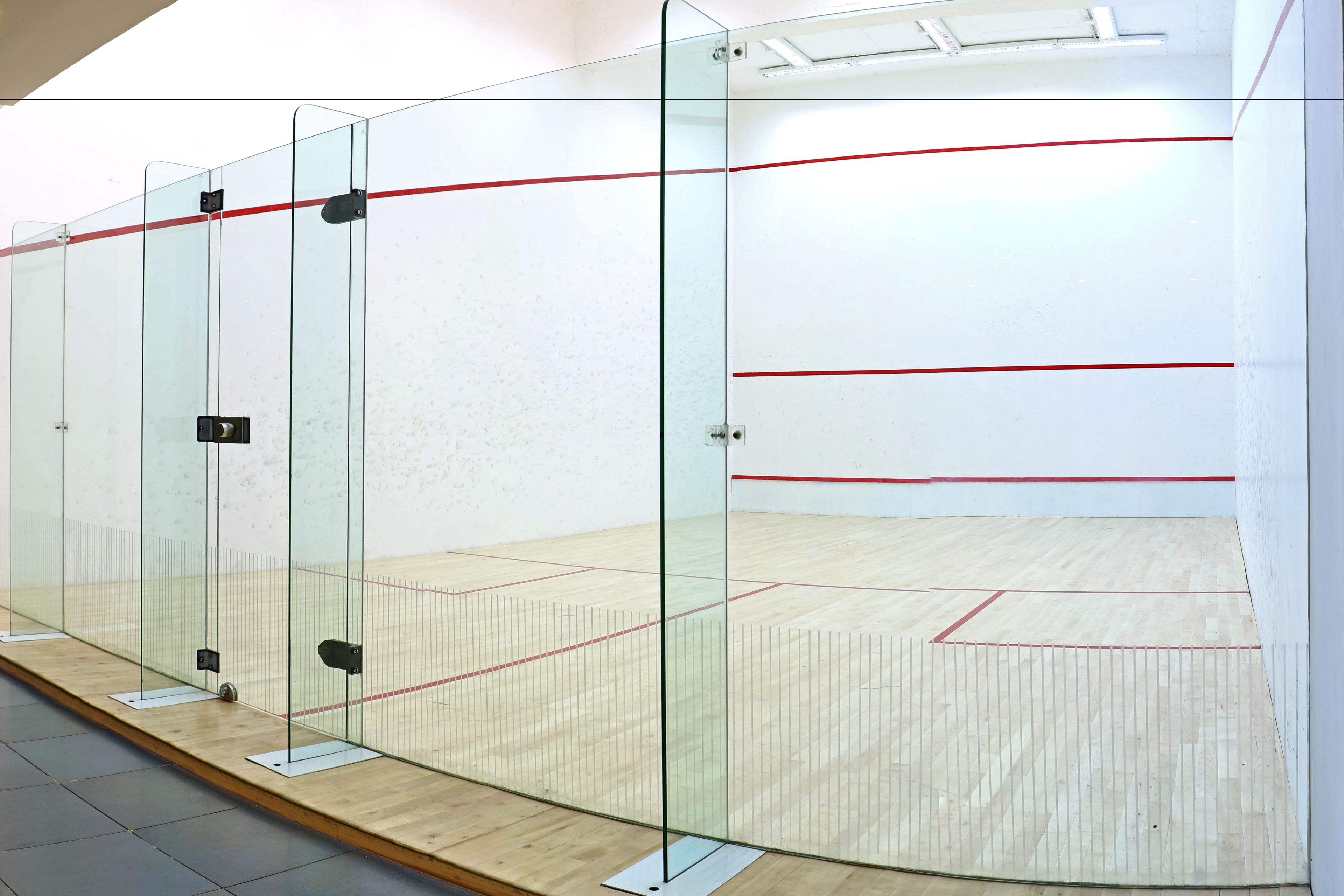 Una cancha de squash con paredes de vidrio, piso de madera y líneas rojas que delimitan el área de juego.