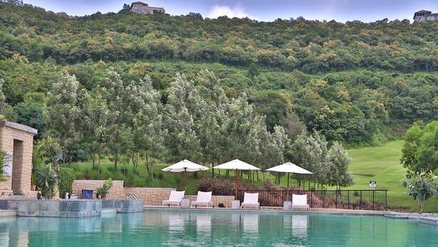 Una piscina con sillas de descanso y sombrillas, rodeada de exuberante vegetación y un campo de golf al fondo.