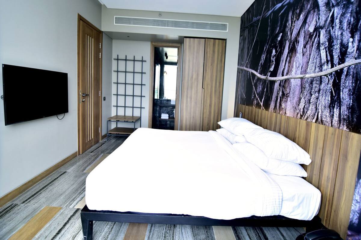 A modern hotel room with a large bed, a flat-screen TV mounted on the wall, a wooden door, and a large abstract painting on the wall.