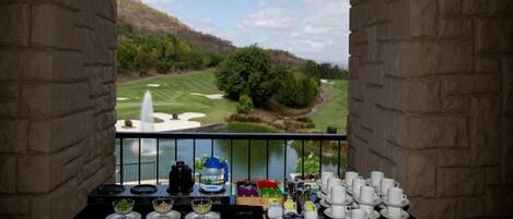 A balcony with a view of a golf course, a fountain, and a table set with cups and a kettle.