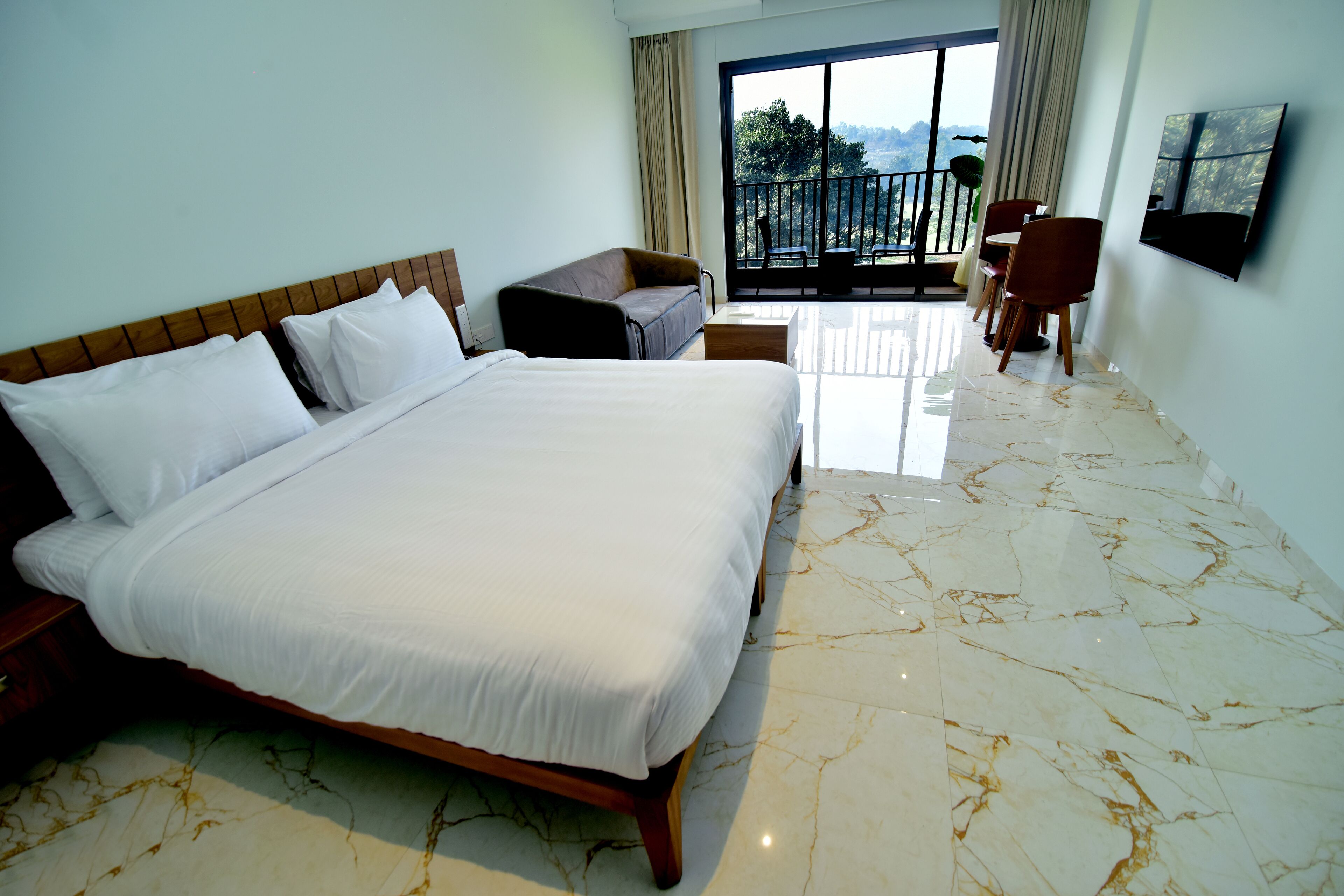 A hotel room with a large bed, a small sofa, a dining table, and a balcony with a view.