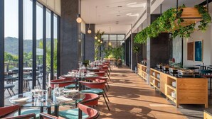 Un moderno comedor con mesas y sillas de vidrio, amplias ventanas que ofrecen vistas al exterior y una encimera de madera con diversos objetos.