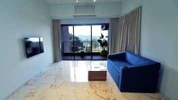 A modern living room with a blue sofa, a flat-screen TV mounted on the wall, and a large sliding glass door leading to a balcony with outdoor furniture.